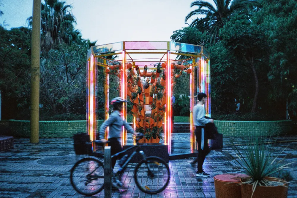 Cyclists pass the glowing BioRhythm pavilion in Grattan Gardens at dusk